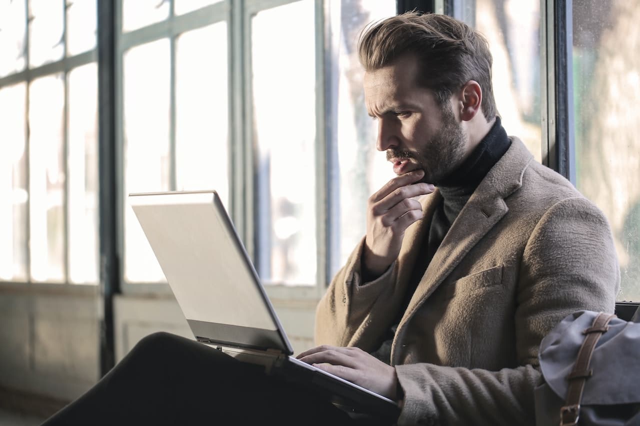 Man in an overcoat sitting by a tall window, hand on his chin, studying an open laptop